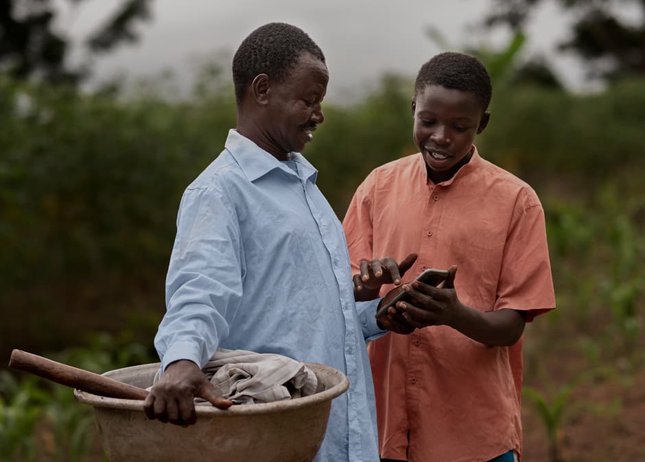 In an agricultural, low-income setting, a man holds farming equipment in one hand and a cellphone in the other. His son shows him how he can earn real rewards on his phone.